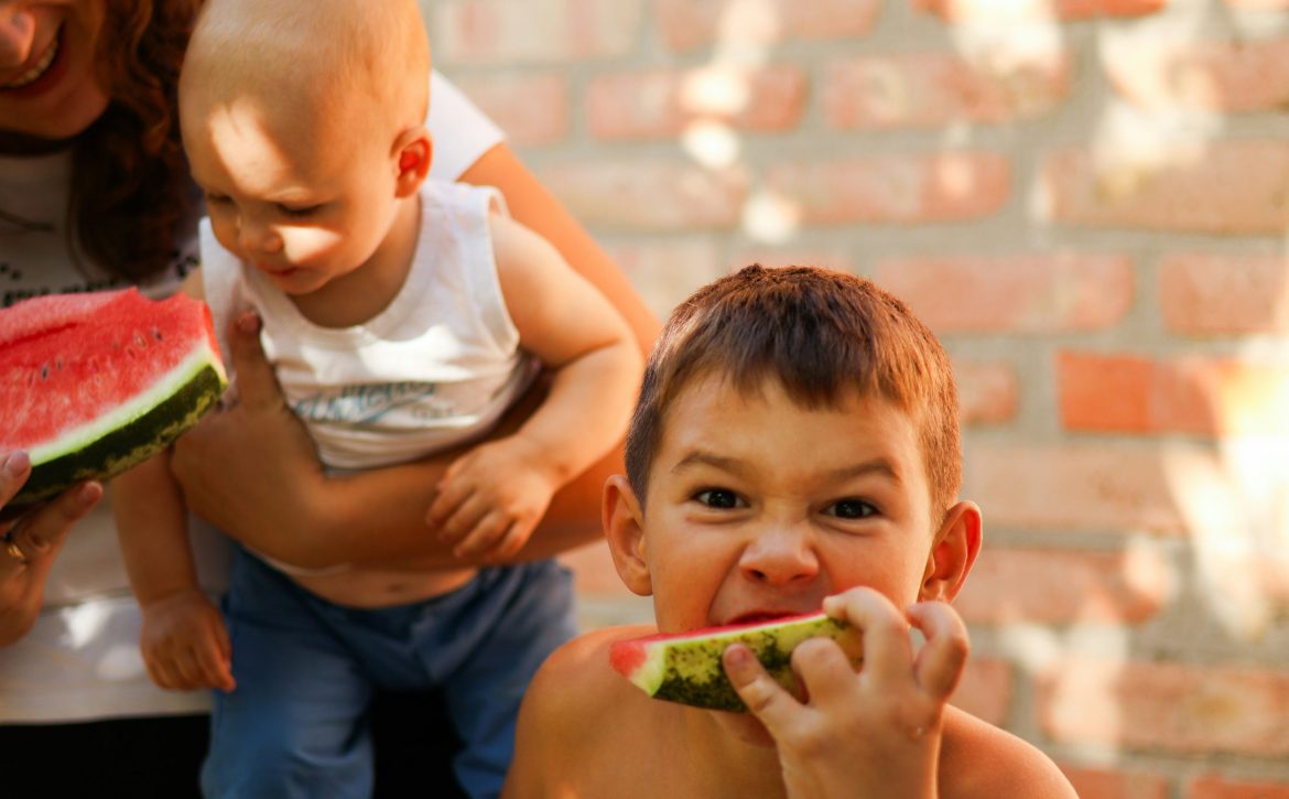 Junge mit Wassermelone in der Hand grinst in Kamera, im Hintergrund ein Baby auf dem Arm der Mutter