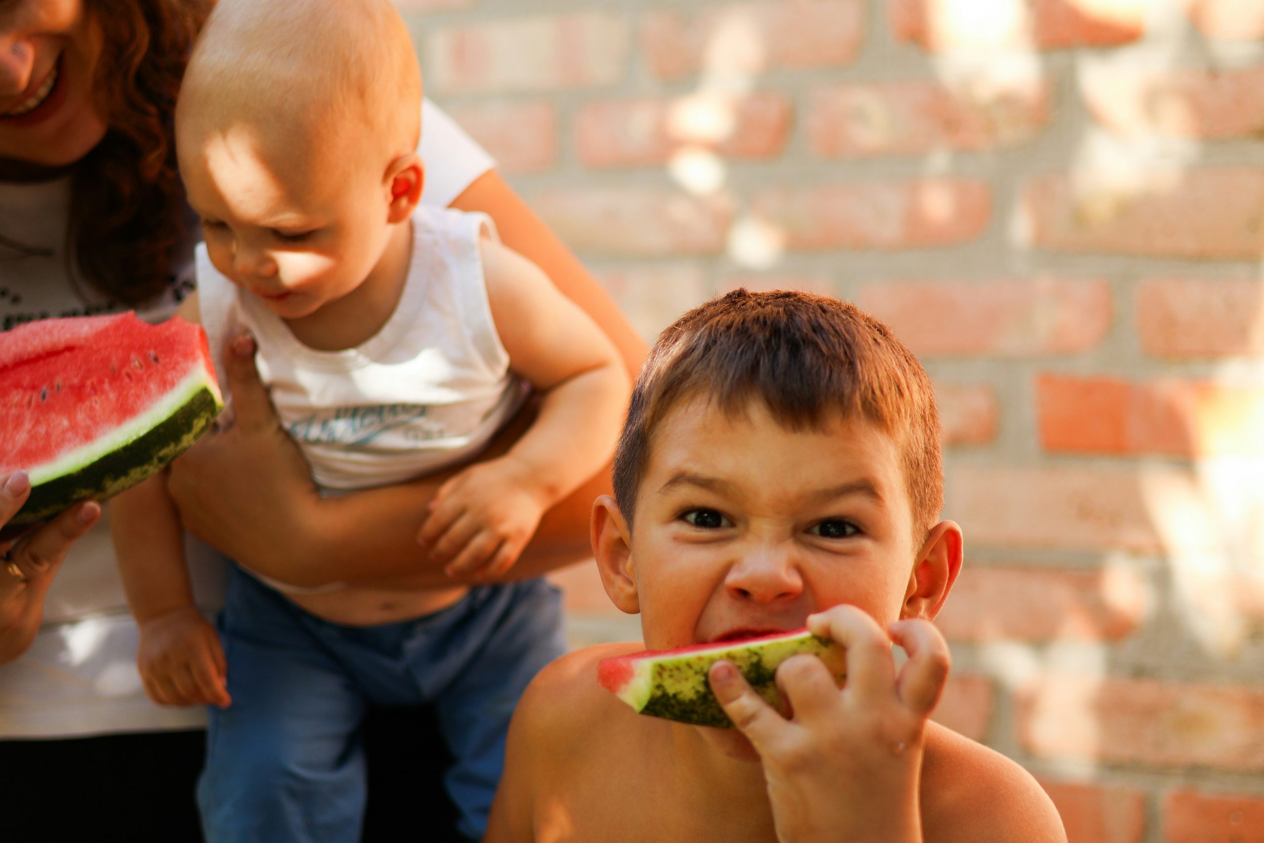 Junge mit Wassermelone in der Hand grinst in Kamera, im Hintergrund ein Baby auf dem Arm der Mutter