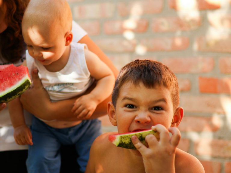 Junge mit Wassermelone in der Hand grinst in Kamera, im Hintergrund ein Baby auf dem Arm der Mutter