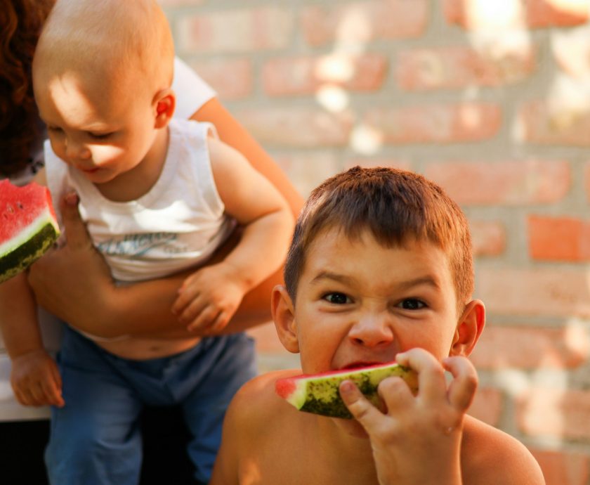 Junge mit Wassermelone in der Hand grinst in Kamera, im Hintergrund ein Baby auf dem Arm der Mutter