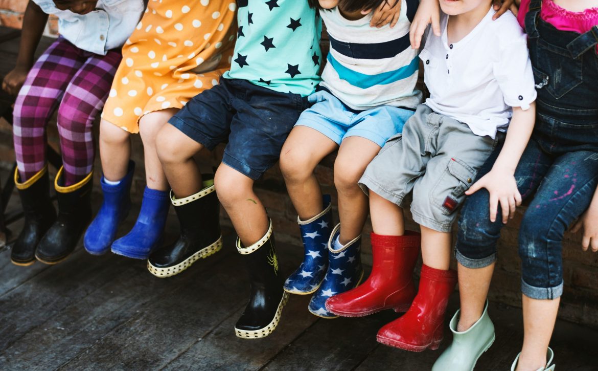 Kinder sitzen auf einer Bank, der Fokus ist vor allem auf ihren Beinen. Alle Kinder tragen bunte Gummistiefel