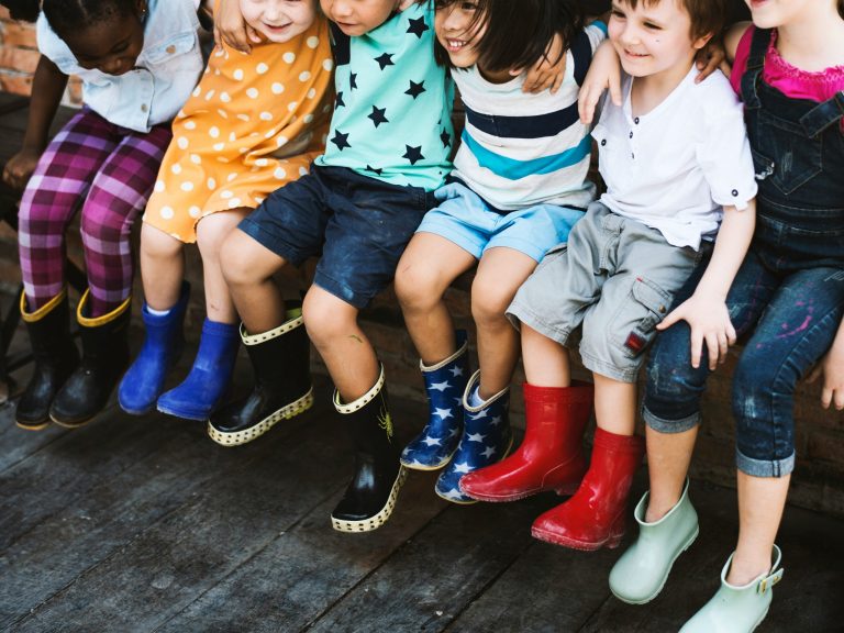 Kinder sitzen auf einer Bank, der Fokus ist vor allem auf ihren Beinen. Alle Kinder tragen bunte Gummistiefel