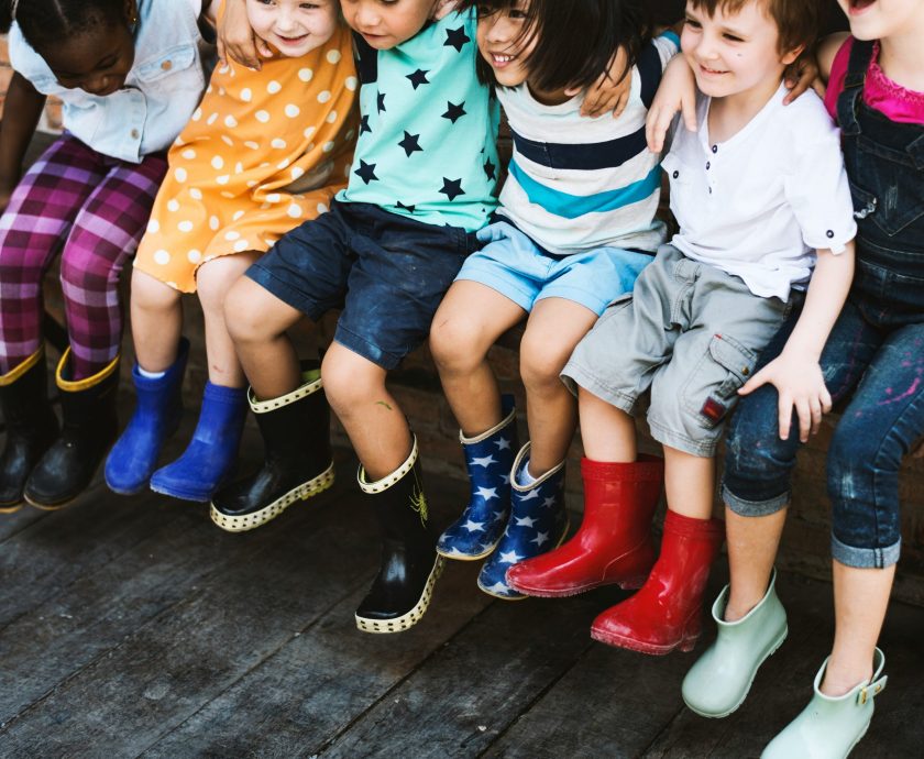 Kinder sitzen auf einer Bank, der Fokus ist vor allem auf ihren Beinen. Alle Kinder tragen bunte Gummistiefel